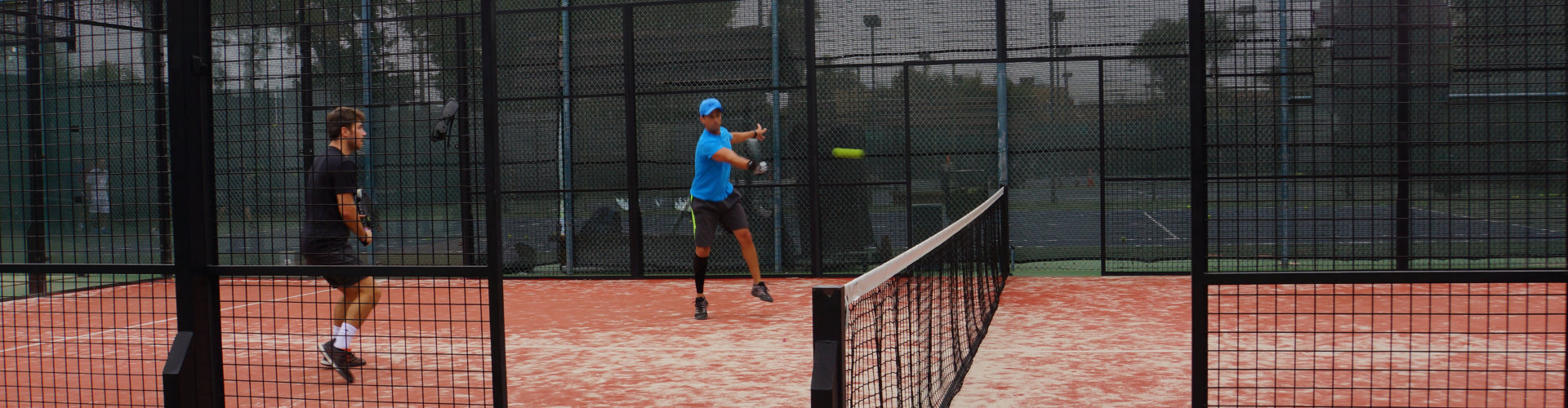 Two players in a padel rally on one of South End's enclosed courts, one hitting a forehand near the net